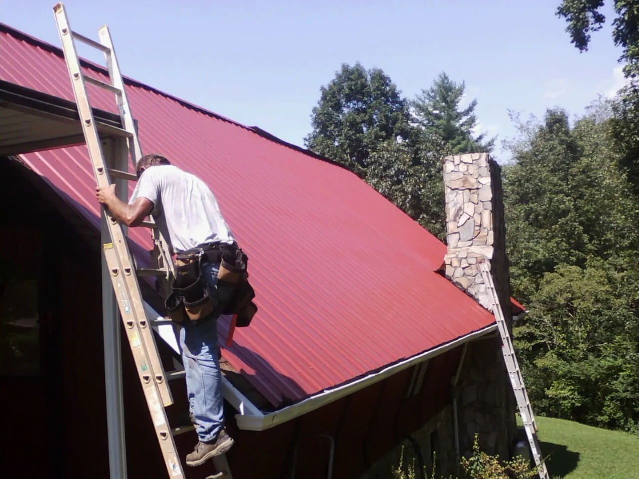 Roofer on a ladder beside a red metal roof and stone chimney — Soffit & Fascia Repair by Roofing Brothers in Eaton Park